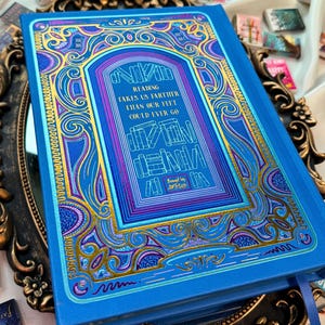 May include: A blue book with gold and purple decorative accents and the text "Reading takes us farther than our feet could ever go." The book is surrounded by other miniature books.