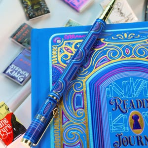 May include: A blue reading journal with gold detailing and a matching pen. The journal features the words "READING JOURNAL" and a keyhole design. The pen is blue with gold accents and rests on the journal. Miniature book replicas are scattered around.