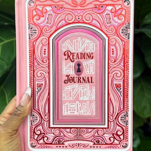May include: A pink "Reading Journal" with decorative cover art. The journal features a keyhole and bookshelf design. The cover is a light pink with darker pink and red accents. The journal is held by a hand.