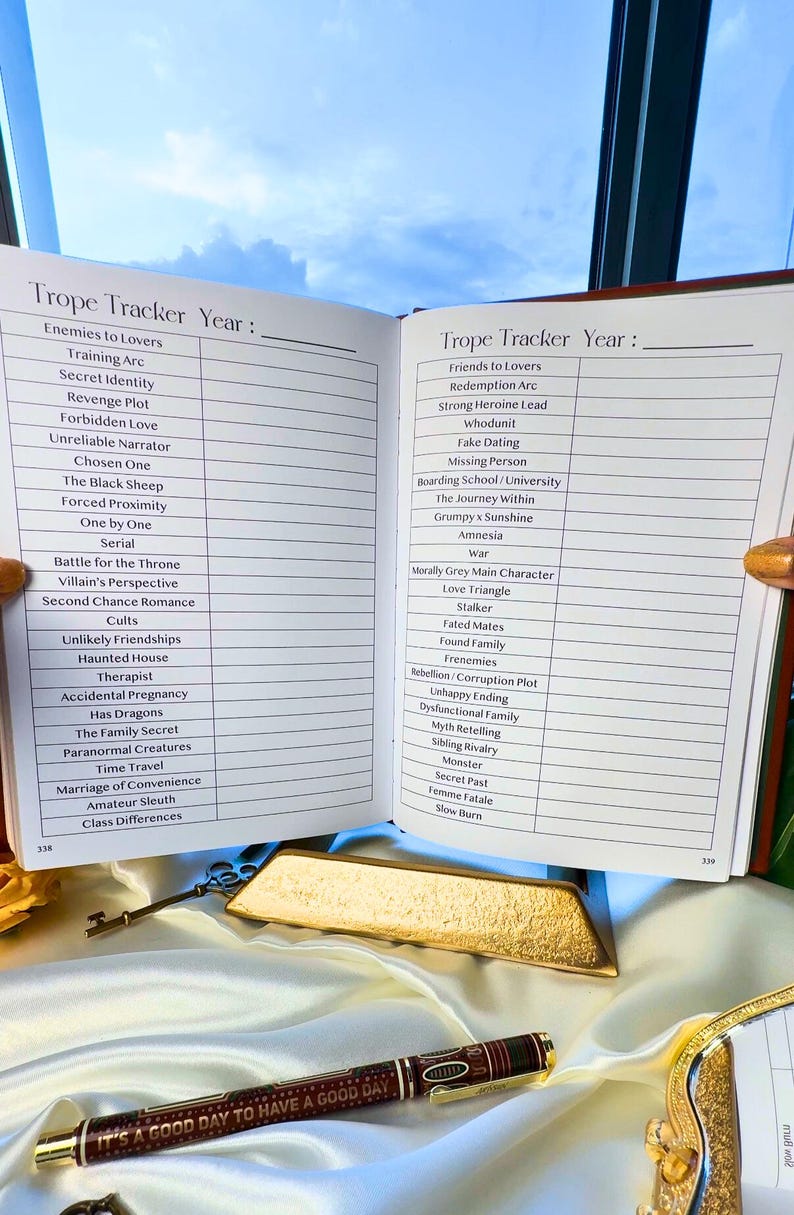 May include: An open notebook titled "Trope Tracker Year" with a list of romance tropes. A pen with the text "It's a good day to have a good day" is on a satin surface.