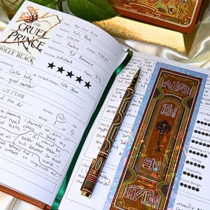May include: An open journal with handwritten notes and a green ribbon. A decorative pen and bookmark with a key design are next to the journal. The book is titled "The Cruel Prince" by Holly Black.