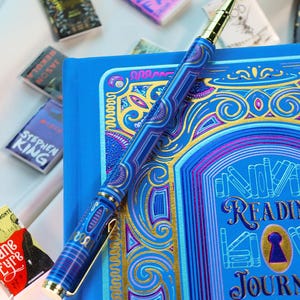 May include: A blue reading journal with gold accents and a matching pen. The pen and journal feature intricate designs in shades of blue and gold. Miniature book-shaped magnets are scattered around the journal.