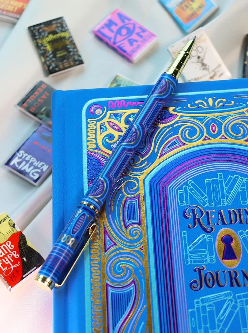 May include: A blue reading journal with gold accents and a matching pen. The pen and journal feature intricate designs in shades of blue and gold. Miniature book-shaped magnets are scattered around the journal.