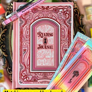 May include: A pink reading journal with an elaborate cover design, featuring the words "READING JOURNAL". A matching pink pen with the text "Buy me books & tell me I'm pretty" and a bookmark are also visible.