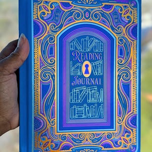May include: A blue "Reading Journal" book with ornate gold and purple detailing. The cover features a keyhole design and the words "Reading Journal" in elegant script. The book is held by a hand.