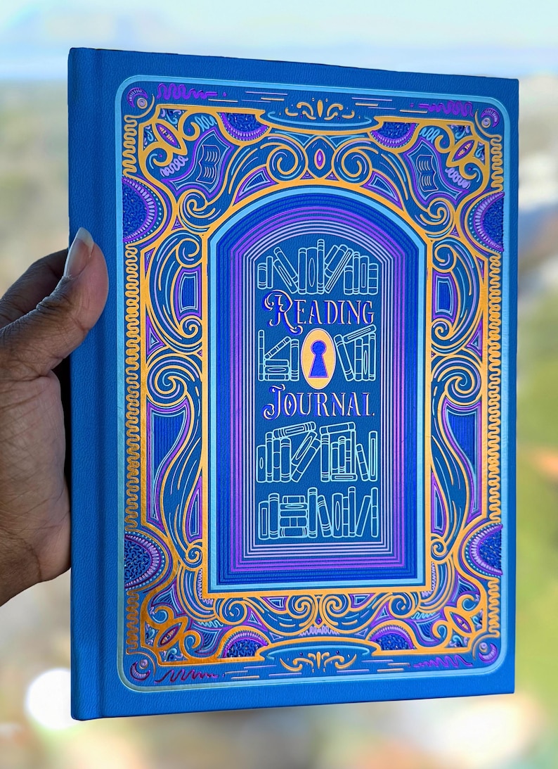 May include: A blue "Reading Journal" book with ornate gold and purple detailing. The cover features a keyhole design and the words "Reading Journal" in elegant script. The book is held by a hand.