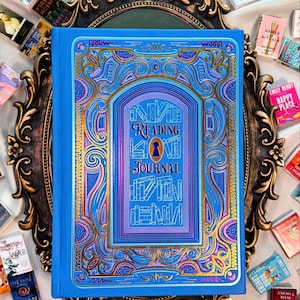 May include: A blue reading journal with an ornate gold and purple design. The cover features the text "Reading Journal" in a decorative font, with a keyhole illustration. The journal is surrounded by miniature books and rests on a decorative, gold-framed mirror.