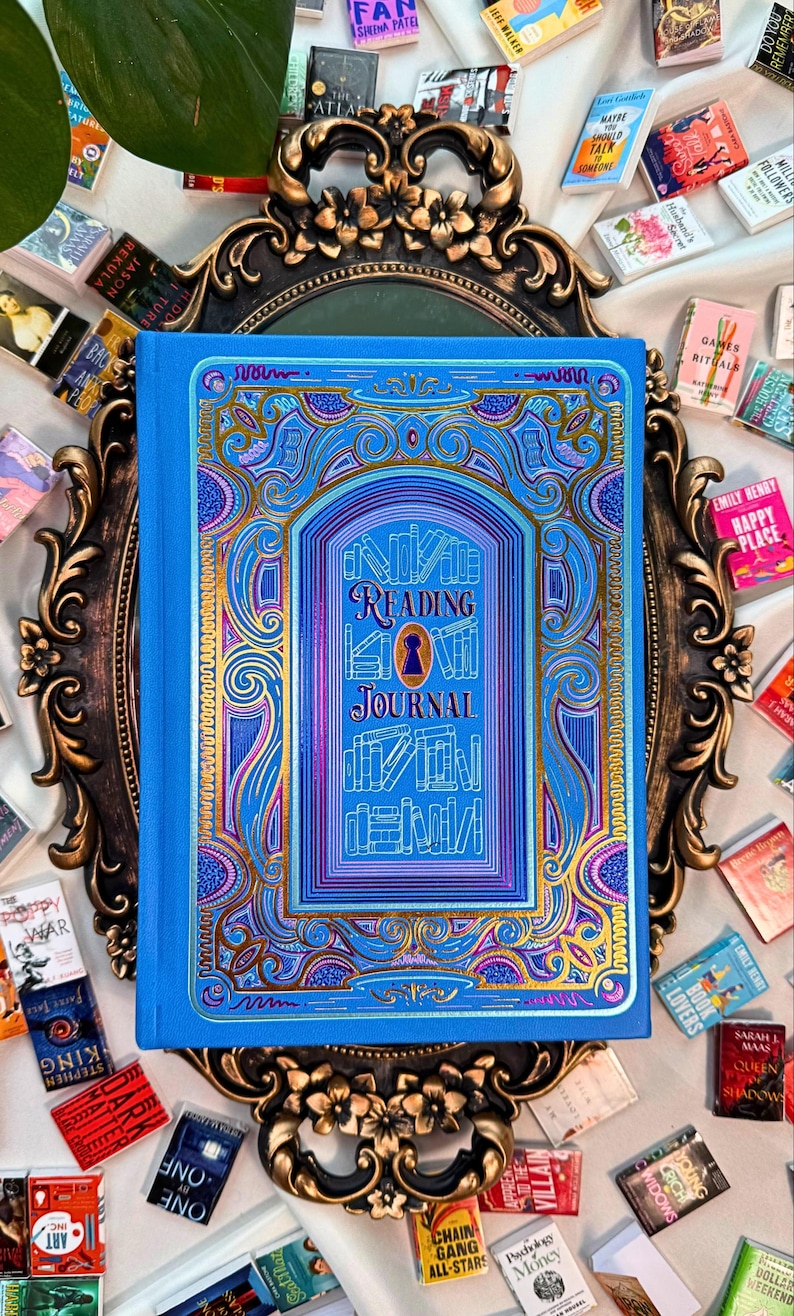 May include: A blue Reading Journal with intricate gold and purple designs, keyhole detail, and the title in elegant lettering. Surrounded by miniature books and set on an ornate, gold-framed mirror, it's a visually rich item for book lovers.