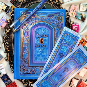 May include: A blue "Reading Journal" with gold and purple detailing, a pen, and miniature books. The journal features a keyhole design and the text "Reading Journal". Two matching bookmarks are also visible.