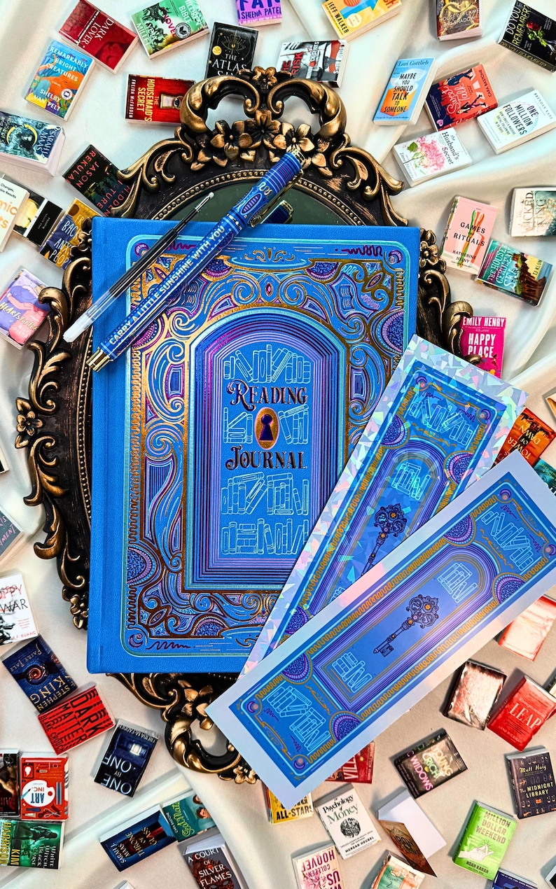 May include: A blue "Reading Journal" with gold and purple accents, a pen, and several miniature books. The journal has a keyhole design and the text "Reading Journal". Two matching bookmarks are also present.
