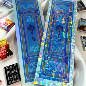 May include: Two holographic bookmarks with a blue door design and a key in the center. The text "NIKA" is visible. The bookmarks are surrounded by miniature books. The text "Pearl Holographic" and "Kaleidoscope Holographic" are also visible.