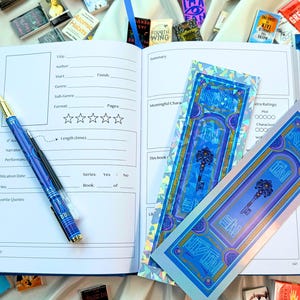 May include: An open blue journal with a pen, and two decorative bookmarks. The pen is blue with gold accents. The bookmarks are blue with a key design. Small books are scattered around the journal.