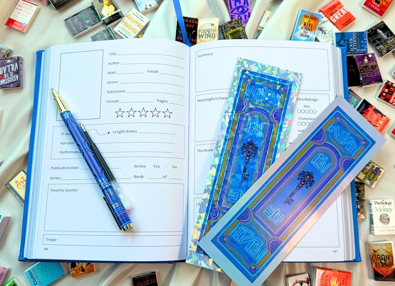 May include: An open blue journal with a pen, and two decorative bookmarks. The pen is blue with gold accents. The bookmarks are blue with a key design. Small books are scattered around the journal.