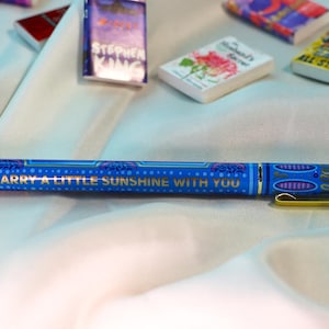 May include: A blue pen with gold accents and the text "CARRY A LITTLE SUNSHINE WITH YOU." Miniature books are scattered around the pen. The pen has a gold clip and tip, and a patterned design.