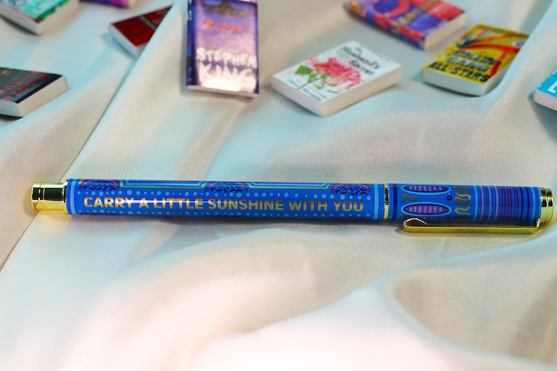 May include: A blue pen with gold accents and the text "CARRY A LITTLE SUNSHINE WITH YOU." Miniature books are scattered around the pen. The pen has a gold clip and tip, and a patterned design.