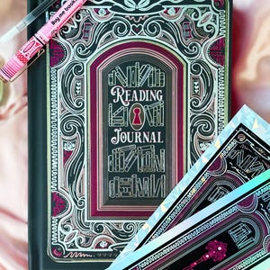 May include: A black Reading Journal with a decorative cover featuring a keyhole design and the words "Reading Journal". A pink pen with the text "Buy me books & tell me I'm pretty" rests on the journal. Two matching stickers are also visible.