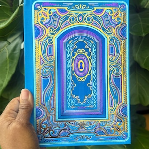 May include: A blue journal with an ornate, layered design featuring gold and purple accents. The cover depicts a keyhole and decorative swirls. The journal is held against a backdrop of green leaves.