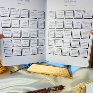 May include: Open book with a 'Book Bingo' game, a pen with the text 'IT'S A GOOD DAY TO HAVE A GOOD DAY', and antique keys. The book has various reading challenges. Gold accents and a white satin surface.