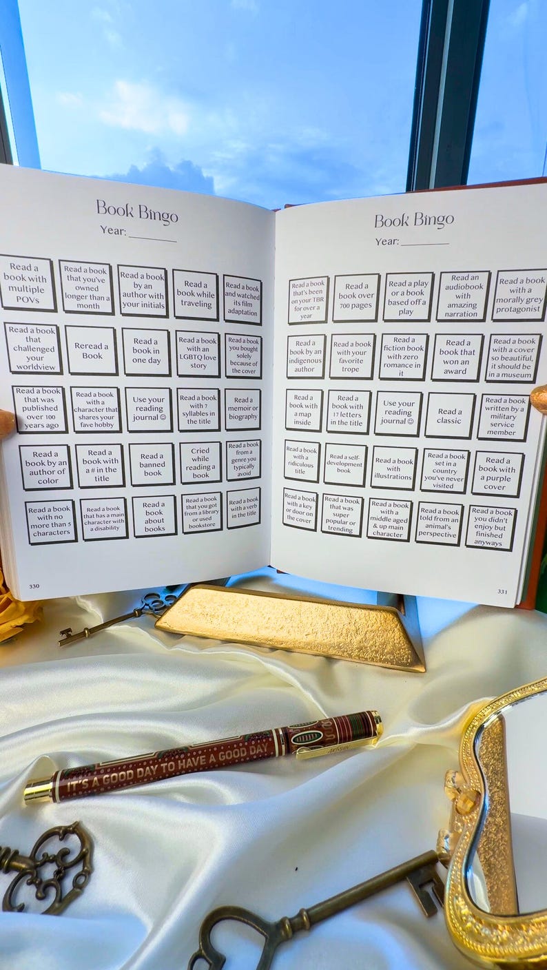 May include: Open book with a 'Book Bingo' game, a pen with the text 'IT'S A GOOD DAY TO HAVE A GOOD DAY', and antique keys. The book has various reading challenges. Gold accents and a white satin surface.