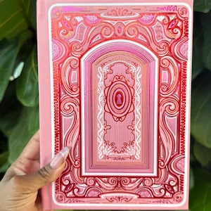 Könnte beinhalten: Rosa Notizbuch mit einem verzierten, mehrschichtigen Design auf dem Einband. Der Einband zeigt ein zentrales Motiv eines gewölbten Türrahmens mit einem Schlüsselloch, umgeben von komplizierten Mustern in Rosa- und Rottönen. Das Notizbuch wird vor einem Hintergrund aus grünen Blättern gehalten.