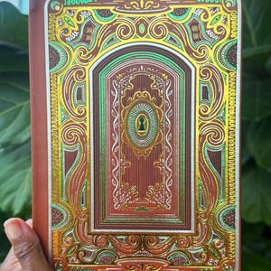 May include: A decorative journal with a brown cover and ornate gold, green, and red embossed design. The cover features a keyhole motif and intricate scrollwork. The journal is held in a hand, with a green leafy background.