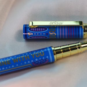 May include: A blue pen with gold accents and the text "BE SUNSHINE WITH YOU." The pen's cap is off, revealing a gold clip with the word "ARTSUN." The pen has a decorative pattern in blue, purple, and gold.