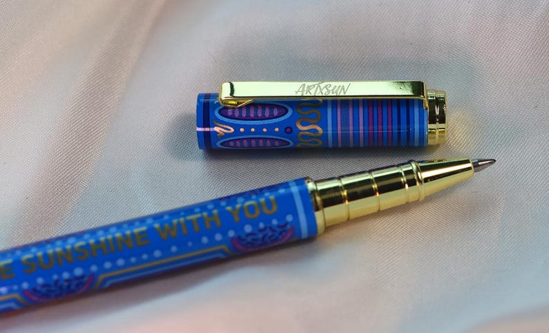 May include: A blue pen with gold accents and the text "BE SUNSHINE WITH YOU." The pen's cap is off, revealing a gold clip with the word "ARTSUN." The pen has a decorative pattern in blue, purple, and gold.