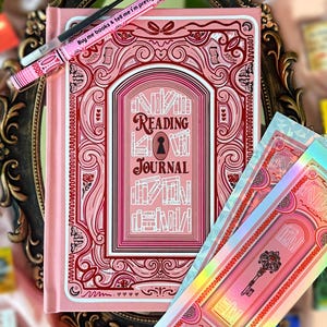 May include: A pink reading journal with a decorative cover featuring the words "Reading Journal" and a keyhole design. A pink pen with the text "Buy me books & tell me I'm pretty" rests on top. Includes matching stickers.