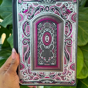 May include: A black journal with a detailed, layered design featuring silver, pink, and black accents. The cover has an ornate, keyhole-shaped central motif. The journal is held against a backdrop of green foliage.