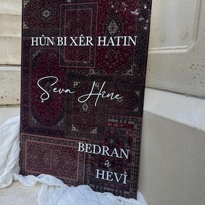 May include: A rectangular sign with a burgundy and brown patterned design, resembling a rug. White text is displayed in a script font and a sans-serif font. The sign is propped up on a stone surface with white fabric draped in front.