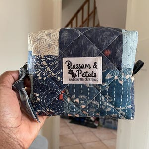 May include: A small, quilted pouch with a patchwork design in shades of blue and cream. The pouch features a diamond-stitched pattern and a label that reads "Blossom & Petals HANDCRAFTED CREATIONS". It has a small loop on the side.