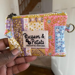 May include: A small, quilted coin purse with a floral patchwork design in pink, purple, blue, and yellow. The purse has a zipper closure, a small loop with a metal ring, and a label that reads "Blossom & Petals HANDCRAFTED CREATIONS."