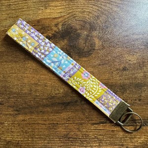 May include: A patterned fabric wrist strap key fob. The design incorporates floral motifs in yellow, purple, and blue. It has a metal ring for keys. The key fob is approximately 15 cm long.