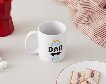 Op de afbeelding: Witte keramische mok met de tekst "Best Dad Ever" in zwart en teal, met een gouden kroon en vlinderdas. De mok staat op een wit oppervlak met een rode espressomaker en een bord met gebak.