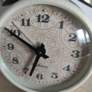 Pode incluir: Um despertador branco vintage com um mostrador creme, ponteiros e algarismos pretos. O mostrador do relógio tem um padrão ornamental e repetitivo em castanho claro. O relógio tem uma campainha prateada no topo. O mostrador tem cerca de 8 cm de diâmetro.