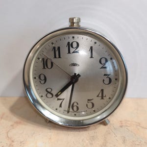 May include: A silver-toned, round analog alarm clock with a light silver face. The clock features black numerals, hands, and the word "PRIM" on the face. The clock has a silver metal frame and two small legs.