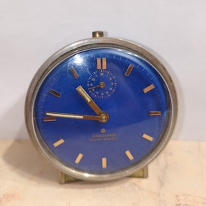 May include: A round analog alarm clock with a vibrant blue face, silver-toned metal casing, and gold-colored hour markers and hands. The dial features the text JUNGHANS BIVOX-ATIC and a small sub-dial for alarm settings at the twelve o'clock position.
