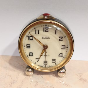 May include: A vintage alarm clock with a cream-colored face, gold-tone bezel, and black casing. The clock face features black numerals, gold hands, and the brand name "SLAVA". A red jewel tops the clock.