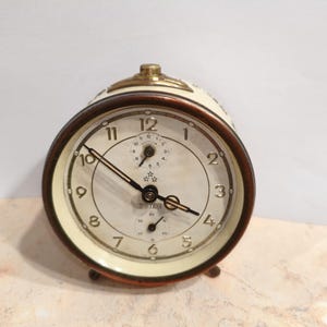 Könnte beinhalten: Ein cremefarbener Wecker im Vintage-Stil mit braunem Rand und goldenen Akzenten. Das Zifferblatt hat schwarze Ziffern und Zeiger sowie ein kleines Sekundenrad. Die Uhr steht auf kleinen Metallfüßen.
