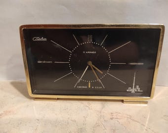May include: Vintage rectangular clock with a black face and gold-colored frame. The clock face features white hour markers and hands, with text in Cyrillic script. The clock has a gold-colored base and is likely from the USSR era.