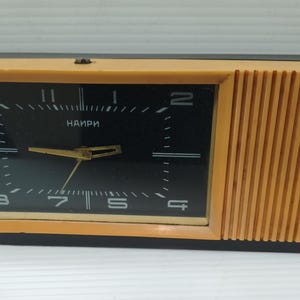 Op de afbeelding: Een rechthoekige vintage klok met een zwarte wijzerplaat en gouden wijzers. De klok heeft een bruin frame en een geribbeld zijpaneel. Het woord "NAIRI" staat op de wijzerplaat.