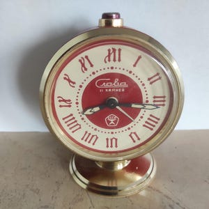Könnte beinhalten: Ein goldfarbener Vintage-Wecker mit rotem und cremefarbenem Zifferblatt. Das Zifferblatt hat römische Ziffern und das kyrillische Wort "Слава". Die Uhr hat ein rundes Zifferblatt und einen kreisförmigen Sockel.