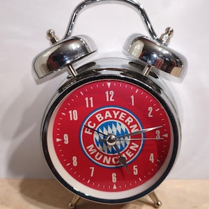 Könnte beinhalten: Ein verchromter Wecker mit rotem Zifferblatt mit dem FC Bayern München Logo. Die Uhr hat eine silberne Glocke und einen Ständer mit drei Beinen. Das Zifferblatt zeigt die Zahlen 1 bis 12.