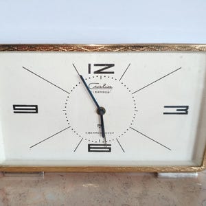 May include: A vintage rectangular clock with a gold-coloured frame. The clock face is off-white with black numerals and hands. The clock face has the text "Слава" and "1 камней" in Cyrillic script. The clock has a gold-coloured base.