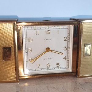 May include: A gold-toned rectangular mantel clock with two hinged side panels featuring a textured finish. The central white clock face displays black Arabic numerals, gold hands, and the text "semca" and "seven jewels" printed in black.