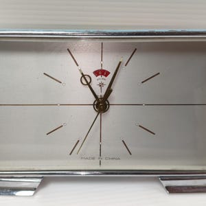 Op de afbeelding: Een rechthoekige vintage klok met een zilverkleurig metalen frame en een lichtgekleurde wijzerplaat. De klok heeft een rood-wit embleem aan de bovenkant en de tekst "MADE IN CHINA" aan de onderkant. De klok is ongeveer 15 cm breed.