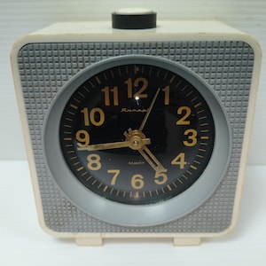 May include: A vintage cream-colored alarm clock with a black face and gold numerals. The clock has a silver-colored ring around the face and a textured gray front panel. The clock has a black button on top and two small feet.