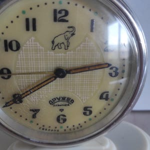Puede incluir: Reloj despertador vintage crema con marco de metal plateado. La esfera del reloj presenta números negros, un elefante y el texto "Дружба". Las manecillas son negras y doradas. El reloj está sobre una base blanca.
