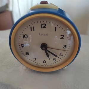 May include: A retro blue and yellow alarm clock with a white face. The clock features black hands and a red button. The clock face displays the word "Rumash" and the text "Made in USSR".