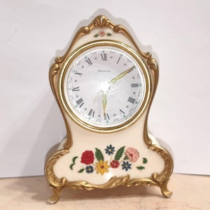 May include: A cream-colored mantel clock with gold-toned ornate trim and a floral motif at the base. The clock face features black Roman numerals, the word Blessing, and gold-toned hands. A small floral accent is also visible at the top peak.